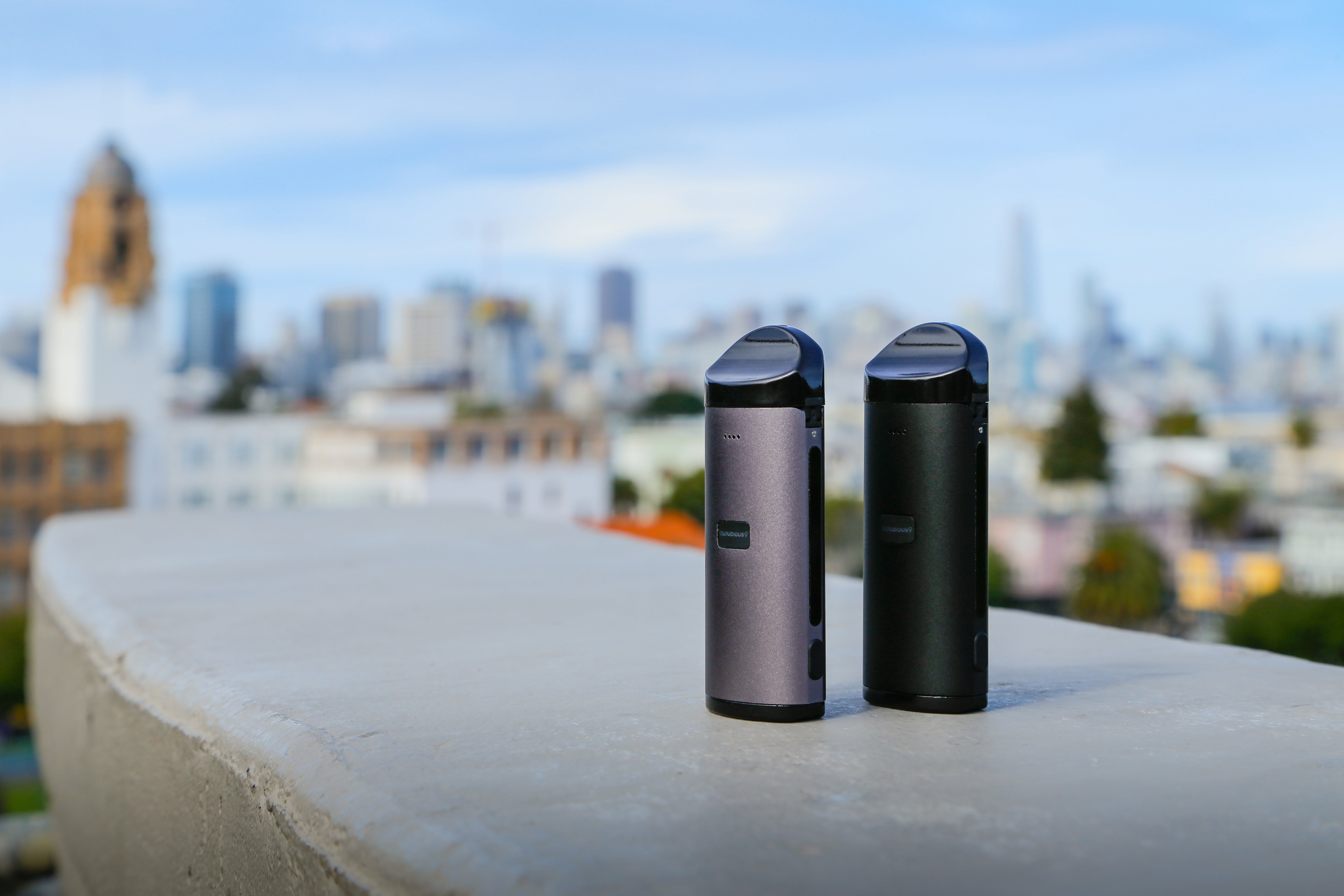 Two Cloudious9 Atomic9 dry flower vapes — one black and one silver — sit perched on a building ledge, overlooking a city skyline. 